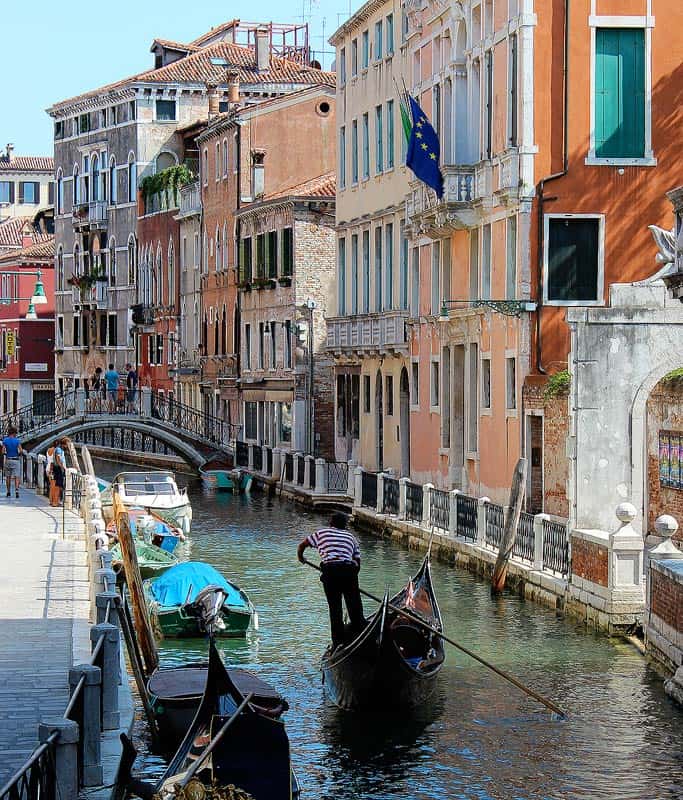 An adorable canal in Venice flanked by colorful Venetian buildings. There is a gondola in the water with a man in black pants and a striped shirt with boats docked along the edge and a bridge in the distance.