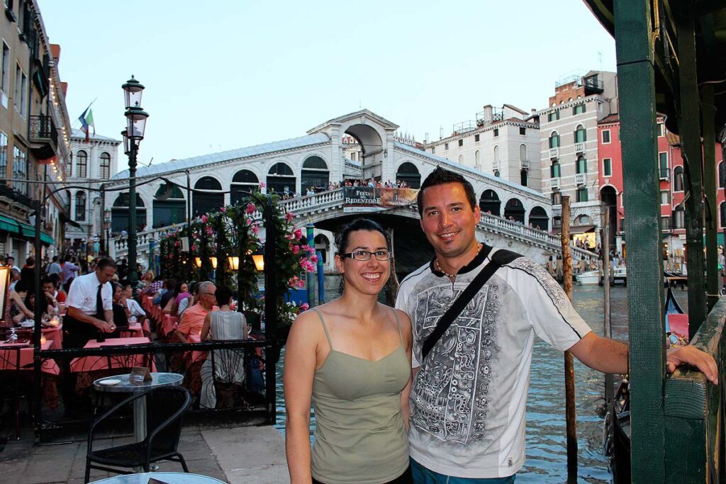 Robyn wearing a light green tank top and Gavin wearing a white and black t-shirt standing in front of Rialto Bridge before enjoying a delicious dinner. There are people eating at tables on the left with green folded umbrellas.