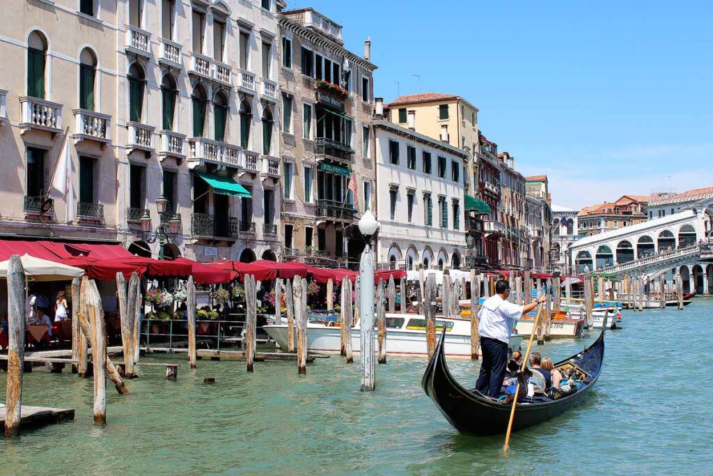 A man in a white shirt steering a gondola on the Grand Canal in Venice in the bottom right of the picture. Classic Venetian buildings are on the left.