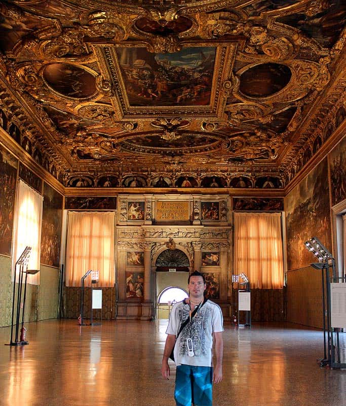 Gavin wearing a white shirt with a black design and blue shorts standing in the middle of a historic palace with ornate gold leaf features and beautiful paintings on the ceiling.