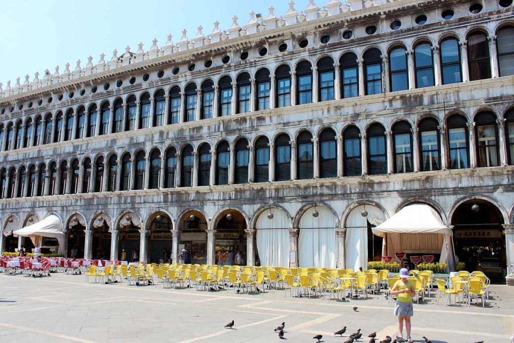 A beautiful Venetian building in St. Mark's square. There are empty round tables and yellow chairs lined up along the sides and a person with pigeons in the bottom right corner.