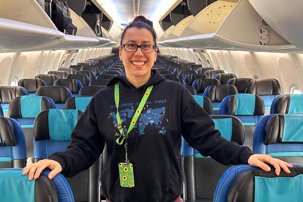 Robyn wearing a black hoodie with a world map smiling in the middle of an empty airplane with black eats and blue headrests. There is a green Sunflower Lanyard around her neck, a signal to airport staff of invisible disabilities.