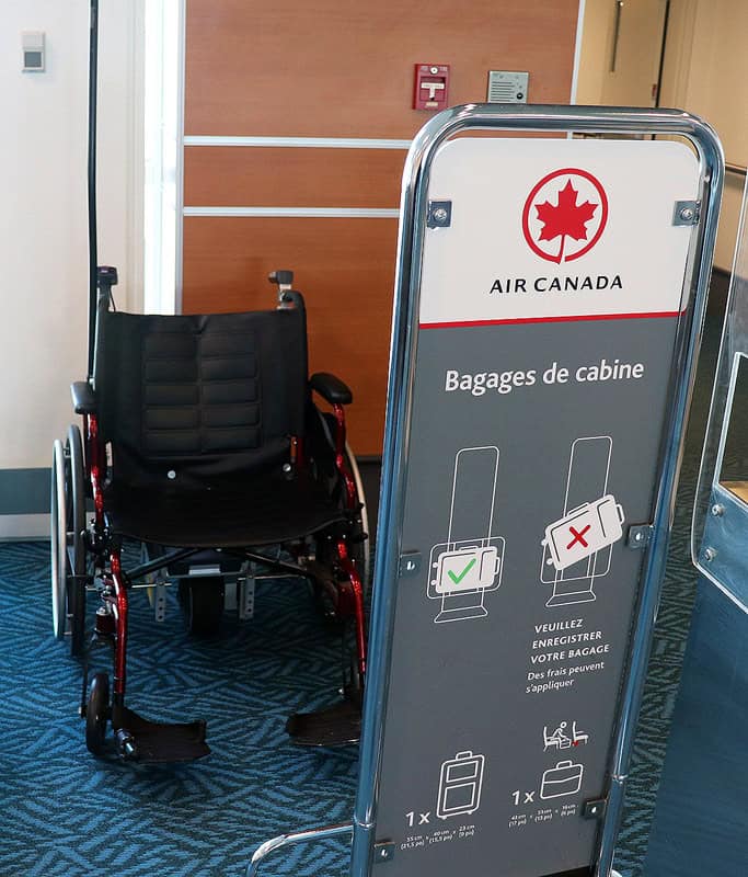 An empty black wheelchair with red accents sitting against the wall beside an Air Canada Baggage Announcement at the gate.