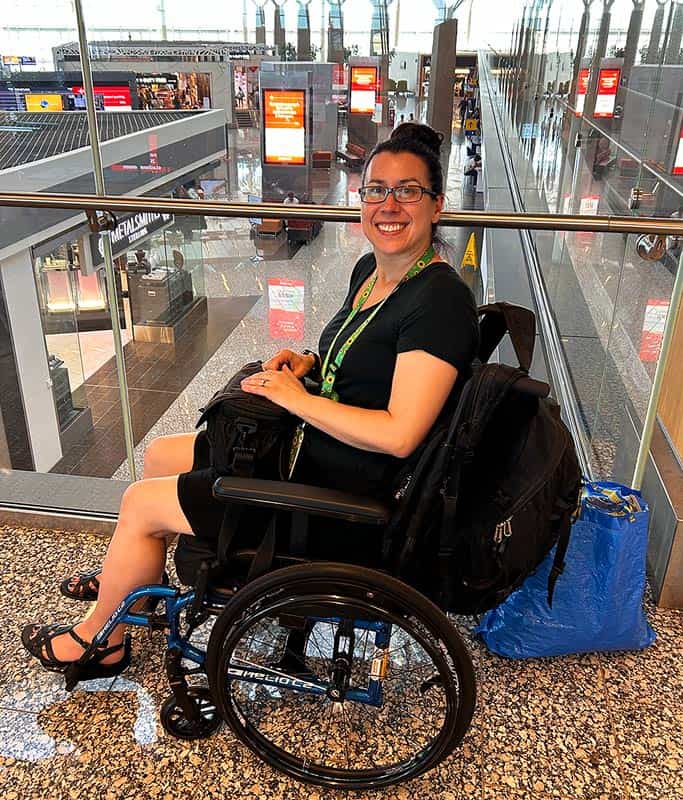 Robyn wearing a black shirt, skirt and Sunflower Lanyard sitting in her wheelchair on a walkway inside the airport. There is a blue bag behind her chair carrying the essentials for her flight.