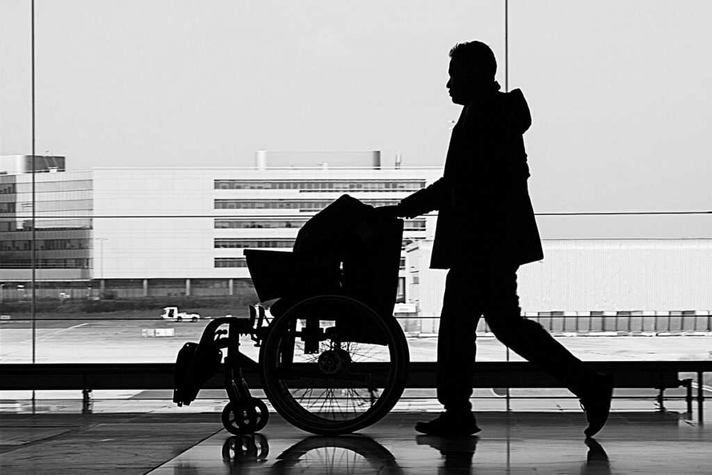 A silhouette of a man pushing a folded up wheelchair through an airport. The image is black and white and the man is walking past floor to ceiling windows.