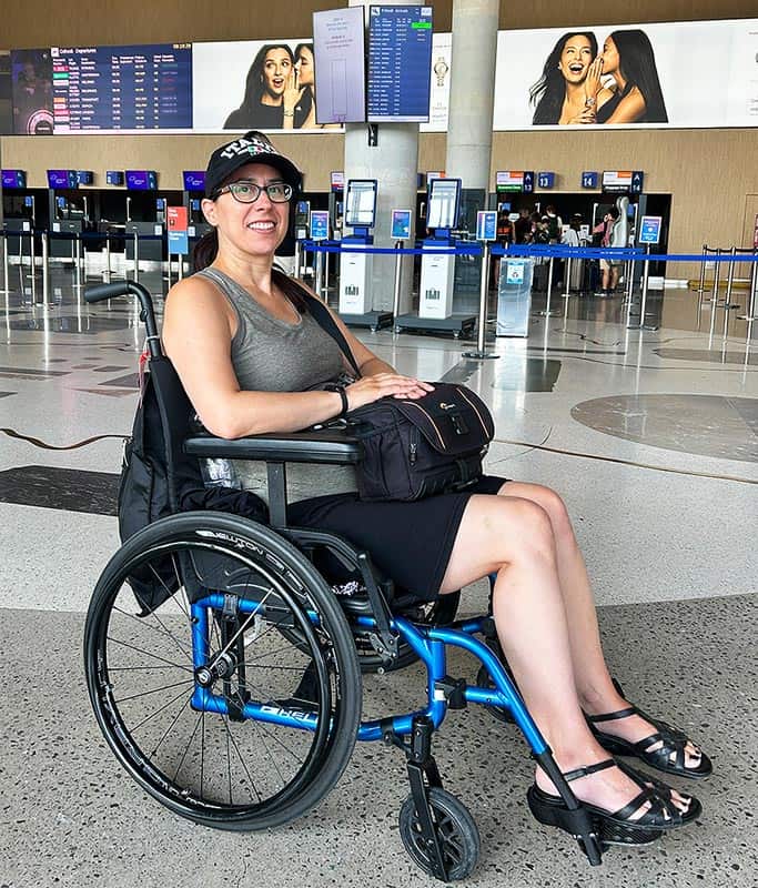 Robyn wearing a green tank top, black skirt and black hat with Italy written on it sitting in her wheelchair at the airport in Slovenia waiting for wheelchair assistance.