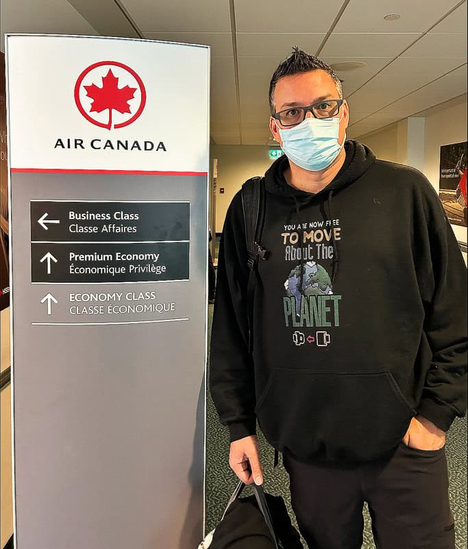 Gavin wearing a face mask and black travel hoodie standing beside an Air Canada sign pointing to Business, Premium and Economy Class doors on his flight. Choosing the right flight can make a big difference when flying with disabilities.