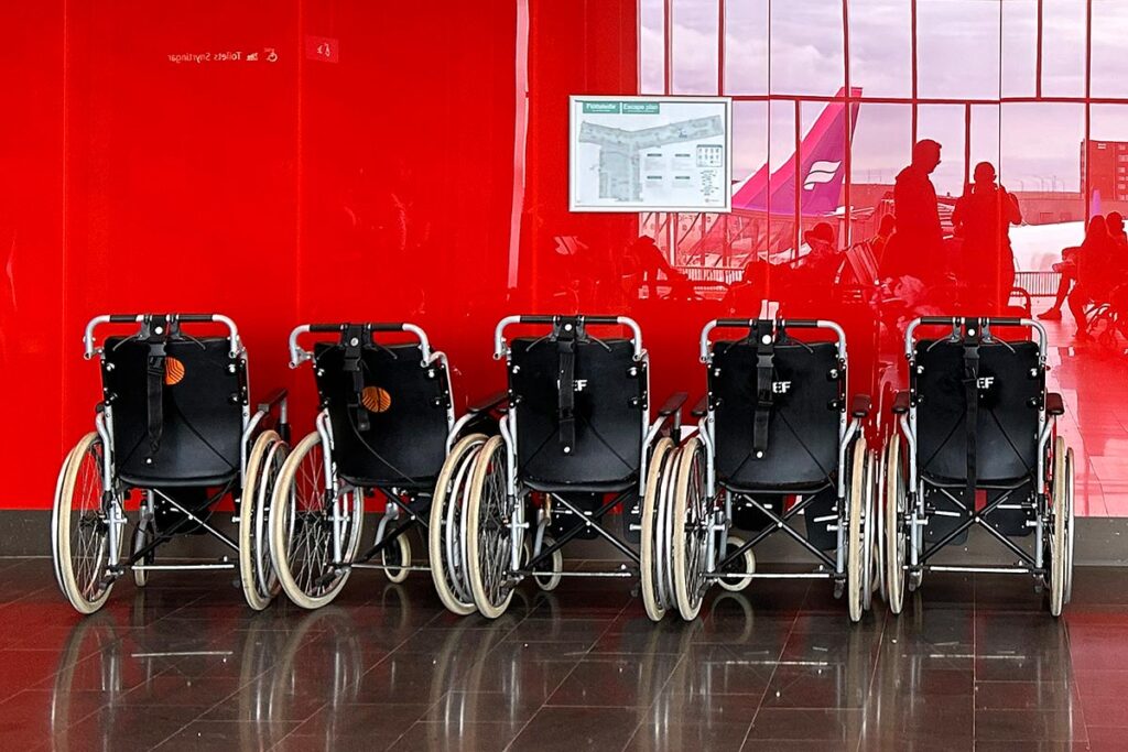 A row of 5 wheelchairs parked against a glossy red wall at the Reykjavik International Airport in Iceland with airplanes in the reflections.