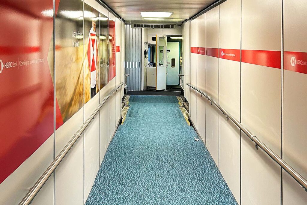 White walls, red advertisements and light blue carpet on the gangway leading to an aircraft. Before the aircraft door is where you will leave your folded wheelchair before boarding.