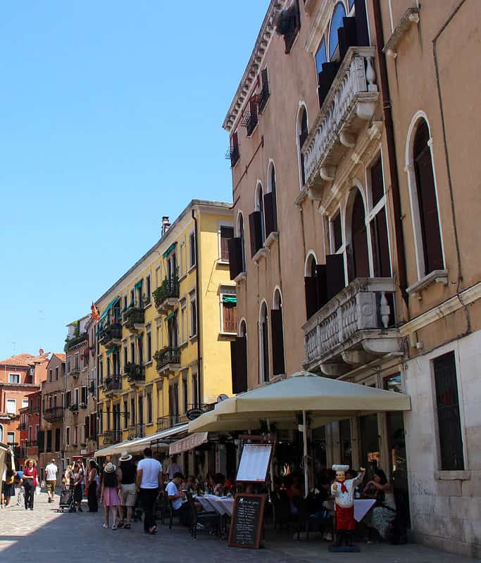 The busy streets of Carraregio, one of the best places to explore in Venice on a day trip. There is a restaurant with outdoor seating in the foreground surrounded by tall, colorful Venetian buildings.