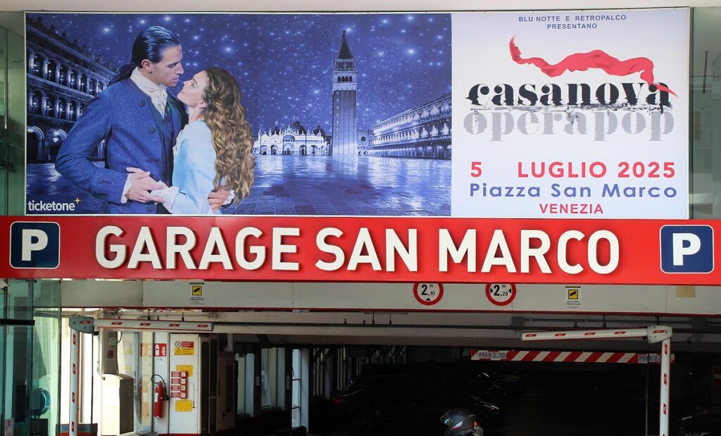 The bright red sign of entrance to San Marco Garage in Venice. There is a large advertisement above showcasing the opera and you can see the dark garage with cars below.