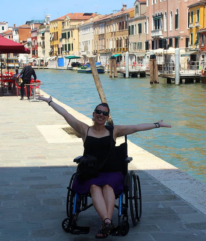 Robyn sitting in her wheelchair wearing a black tank top and purple skirt. Her arms are out and she is next to a canal during her day trip to Venice. There are colorful buildings on the other side of the canal and a red patio umbrella in the background.
