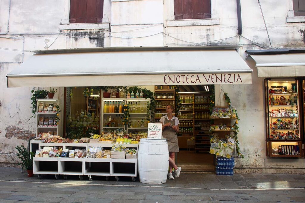 A shop owner wearing a khaki colored apron and white shirt standing beside her outdoor displays of soaps, spices and liquor at the Enoteca Venezia shop in Carraregio area.