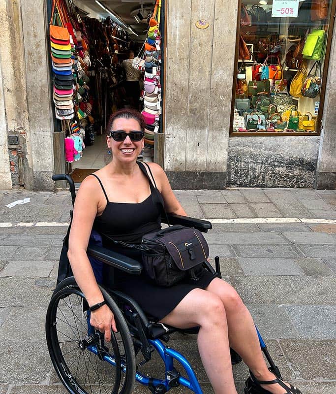 Robyn wearing a black tank top and sunglasses, sitting in her wheelchair smiling in front of the best place to buy Italian leather purses in Venice. Colorful purses line the doorway inside.
