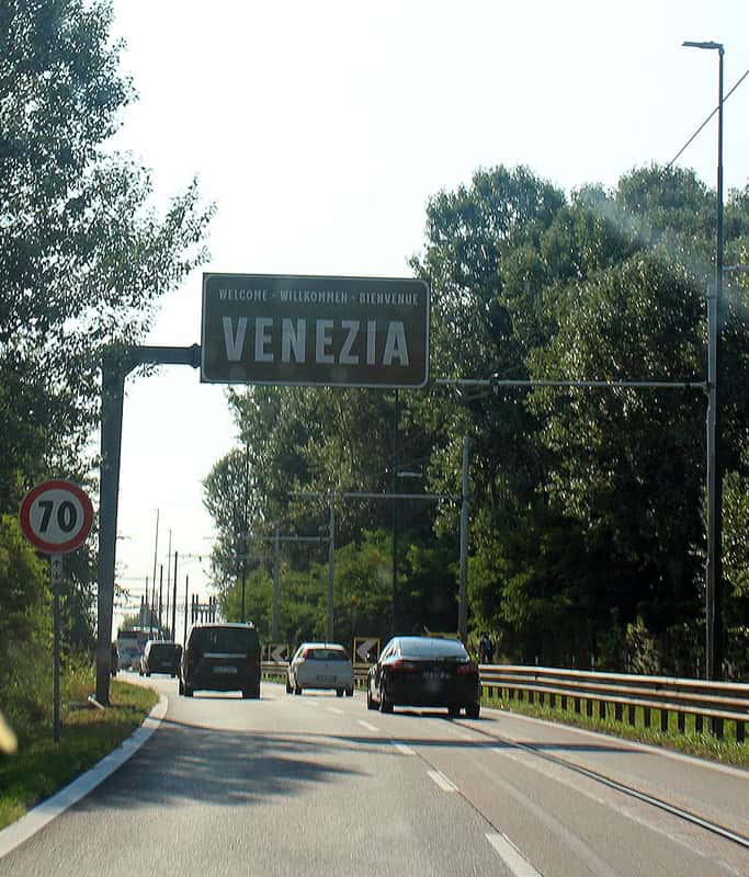 The dark green sign for Venezia above the highway driving to Venice. There are several cars ahead with trees on either side and a 70 speed sign on the left.
