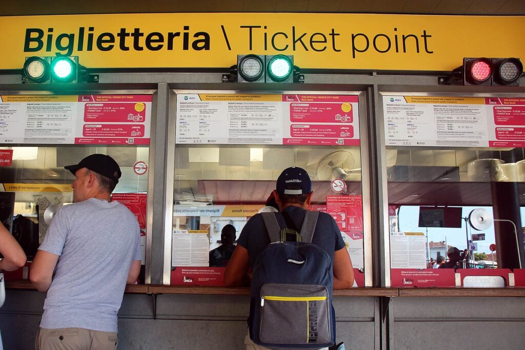 The Vaporetto ticket booth with bright yellow sign outside of the Santa Lucia train station in Venice. Two men in ball caps are at the ticket booths purchases passes.