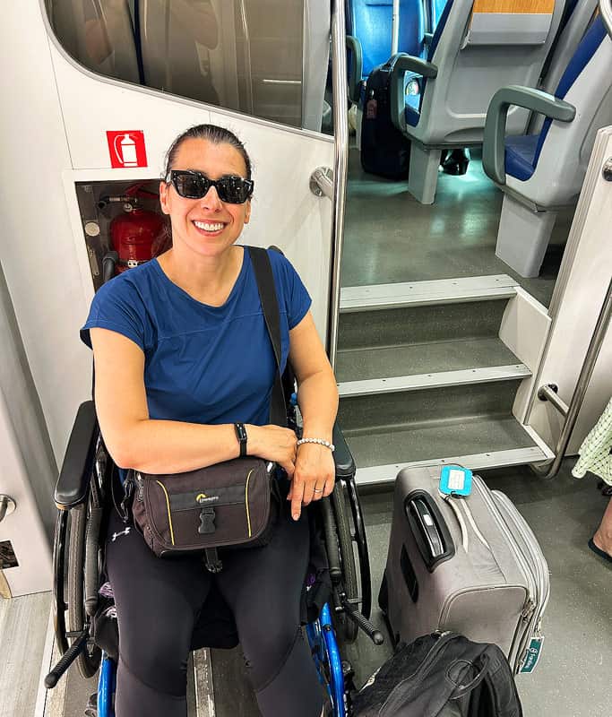 Robyn smiling in sunglasses, wearing a blue t-shirt and black tights sitting in her wheelchair holding a camera bag in the train car going for a Venice day trip. There are stairs up to the seating area in the upper right.