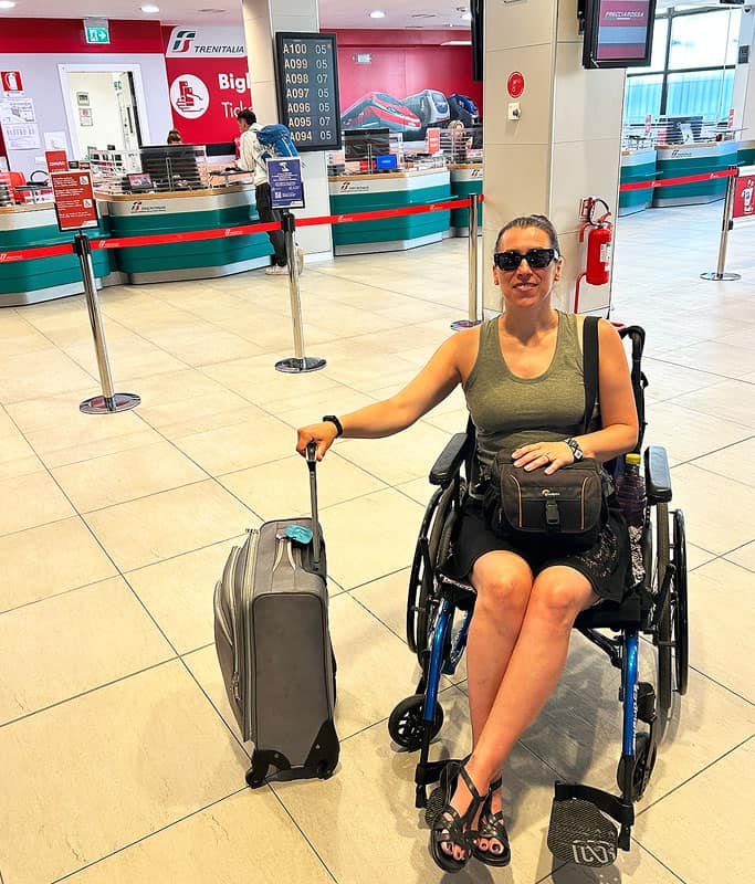 Robyn wearing a green tank top, black skirt and sunglasses sitting in her wheelchair with a camera bag on her lap and a silver carry-on suitcase next to her inside the Train Italia buying tickets for her day trip to Venice.