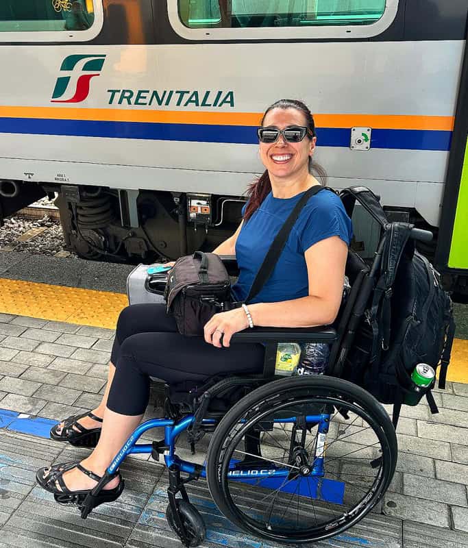 Robyn smiling in sunglasses, wearing a blue tshirt and black tights sitting in her wheelchair holding a camera bag in front of the TrenItalia train car on the platform. There is a black backpack attached to the back of her wheelchair and a silver carry on behind her.