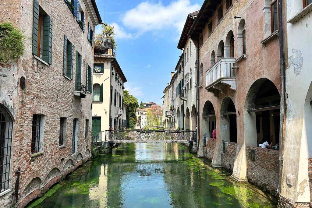 A quiet canal in Treviso with classic brick buildings on ether side and a iron walk bridge in the background. Treviso is an excellent base for a Venice day trip.