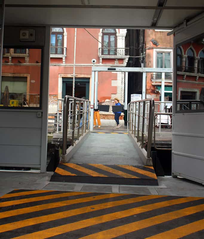 A long ramp with black and yellow stripes connecting the Vaporetto platform to the canal sidewalk in Venice with a person and red building in the background.
