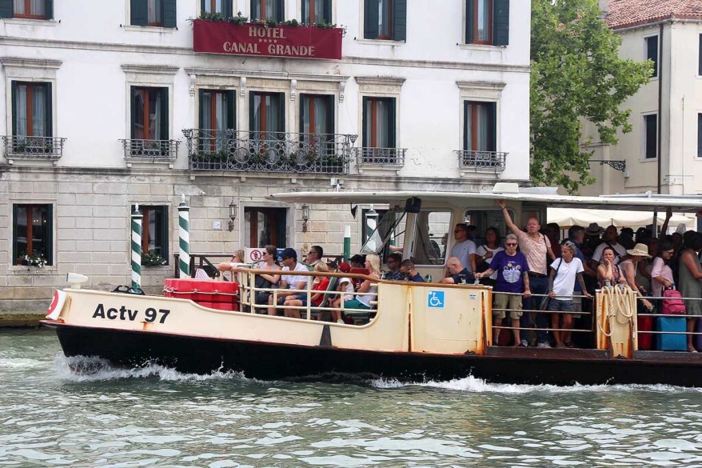 The front half of a Vaporetto in Venice in front of the Grand Canal Hotel. People are packed in the standing area and the front seating area is full.