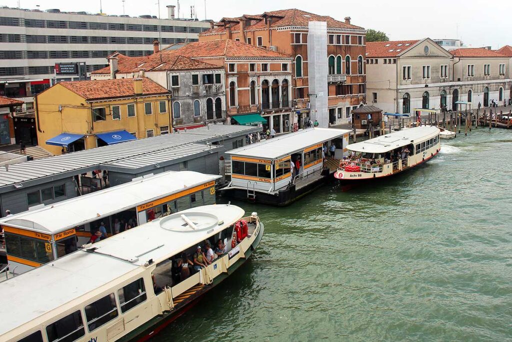 Two Vaporetto stations just outside of the Santa Lucia train station in Venice. There is a vaporetto parked in front of each station, letting people on and off the water bus. There are colorful buildings in the background.