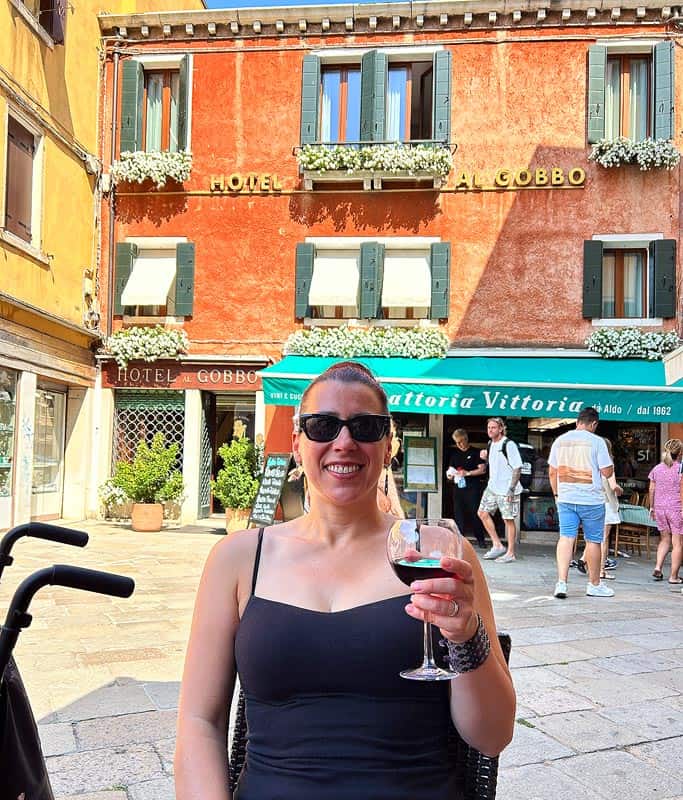 Robyn wearing a black tank top and sunglasses holding a glass of red wine at a restaurant in Carraregio. There is a bright orange building with green shutters behind her.