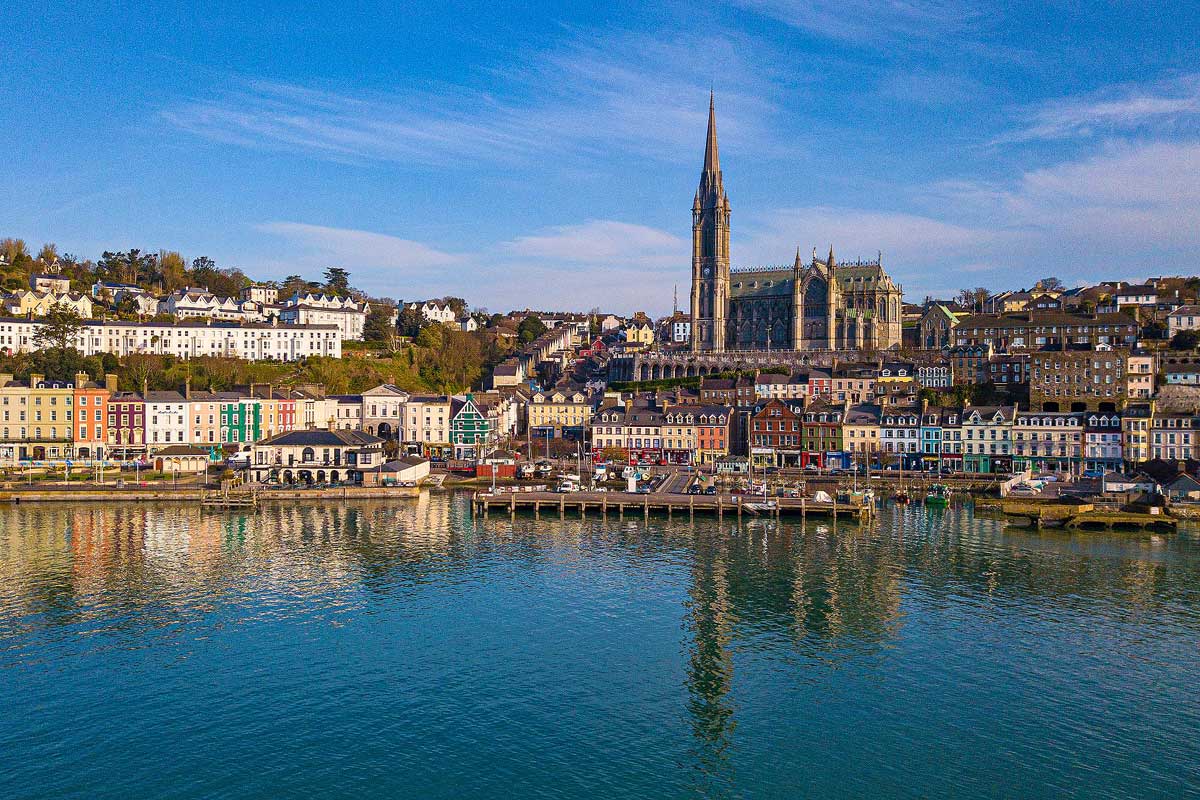The beautiful seaside town of Cobh, a highlight on this wheelchair accessible tour of Ireland. The town is full of colorful buildings and a very tall cathedral in the distance, all perfectly reflected in the still water below with a blue sky above.