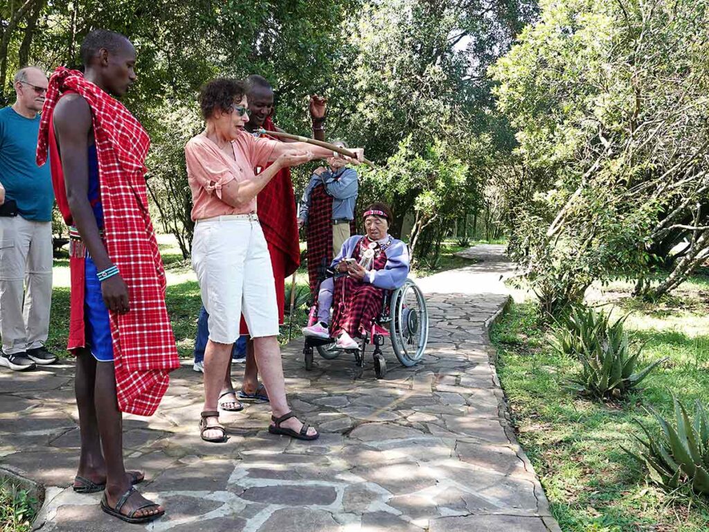 Linda wearing a peach shirt and white shorts using a bow and arrow from the Masai tribe at Naserian Camp. There are two Masai warriors there and a wheelchair user behind her with greenery around.