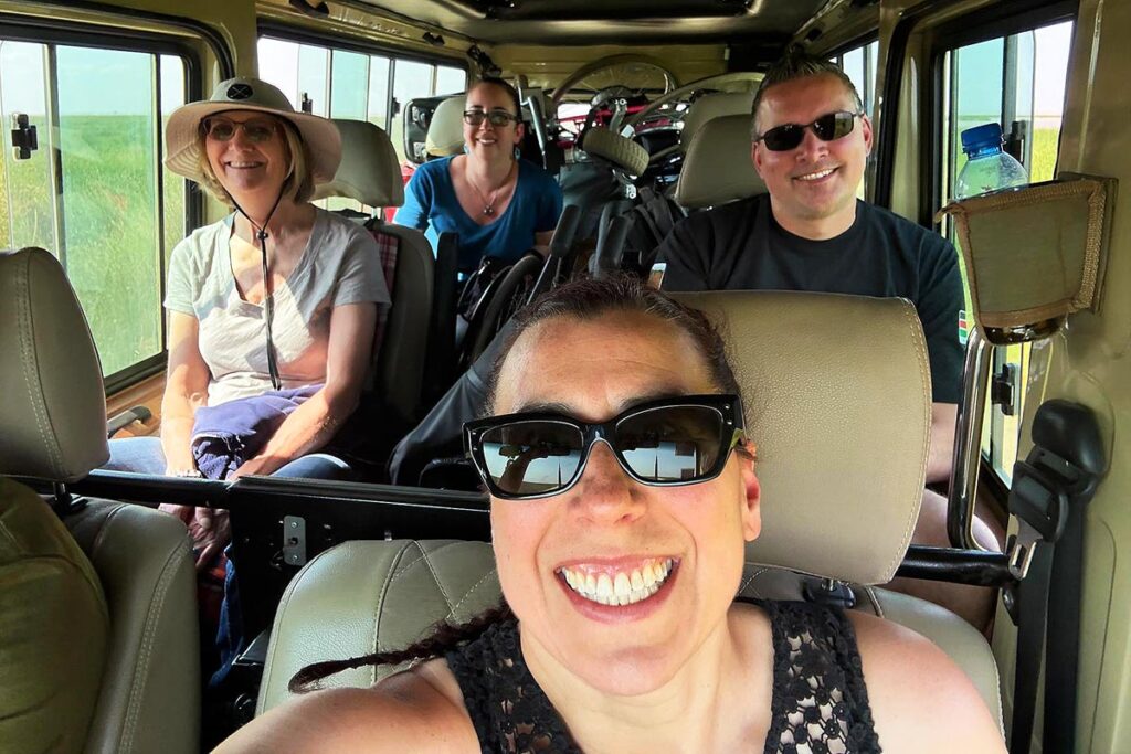 Robyn taking a selfie of her safari jeep in Kenya with Ruth sitting behind on the left, Gavin sitting behind on the right and her sister sitting in the back next to the pile of wheelchairs.