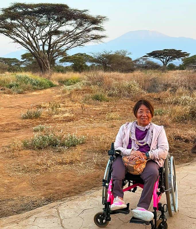 Atsuko sitting in her pink wheelchair wearing a light pink sweater and purple pants smiling in front of Mount Kilimanjaro in Amboseli National Park on her wheelchair accessible Kenya safari in July 2026.