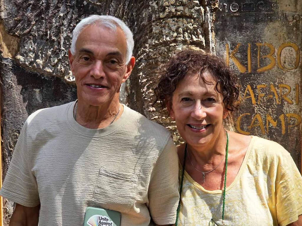 Bob and Linda standing in front of the entry sign at Kibo Safari Camp in Amboseli with an elephant painted on the wood. They are smiling and wearing a grey shirt and yellow shirt respectively.
