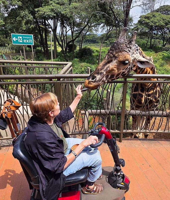 Mary wearing a black long sleeve jacket sitting in her red and black scooter hand feeding a giraffe on the accessible platform at the Giraffe Center in Nairobi with trees in the background.