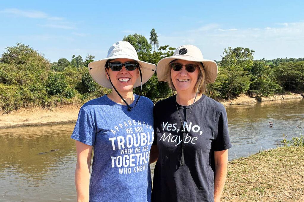 Robyn and Ruth wearing beige safari hats, t-shirts and sunglasses smiling in front of a river full of hippos in Masai Mara, a highlight of our accessible Kenya safari.