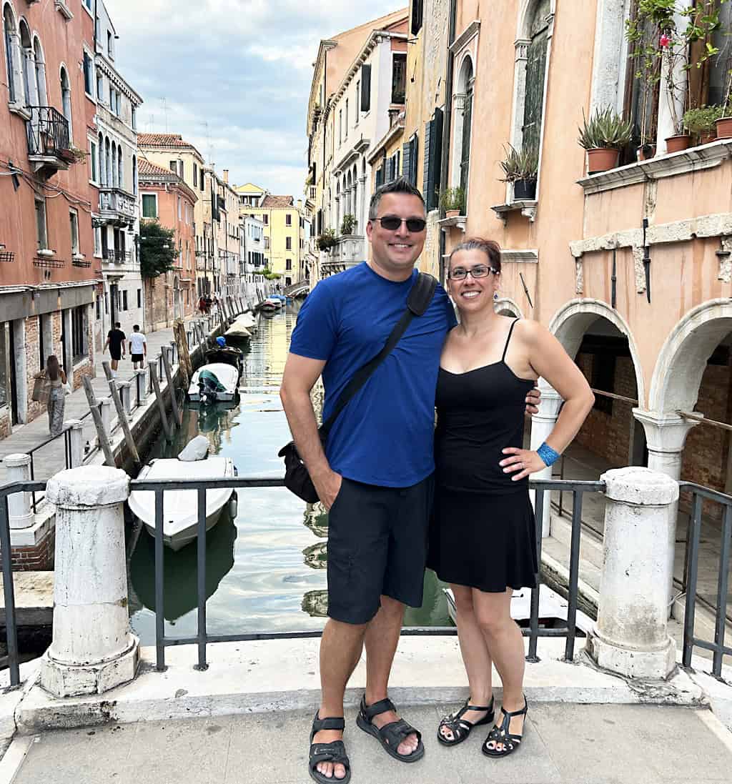 Gavin and Robyn smiling standing together on a foot bridge in Venice. Gavin is wearing a blue shirt and black shorts and Robyn in in a black dress with her hair up.